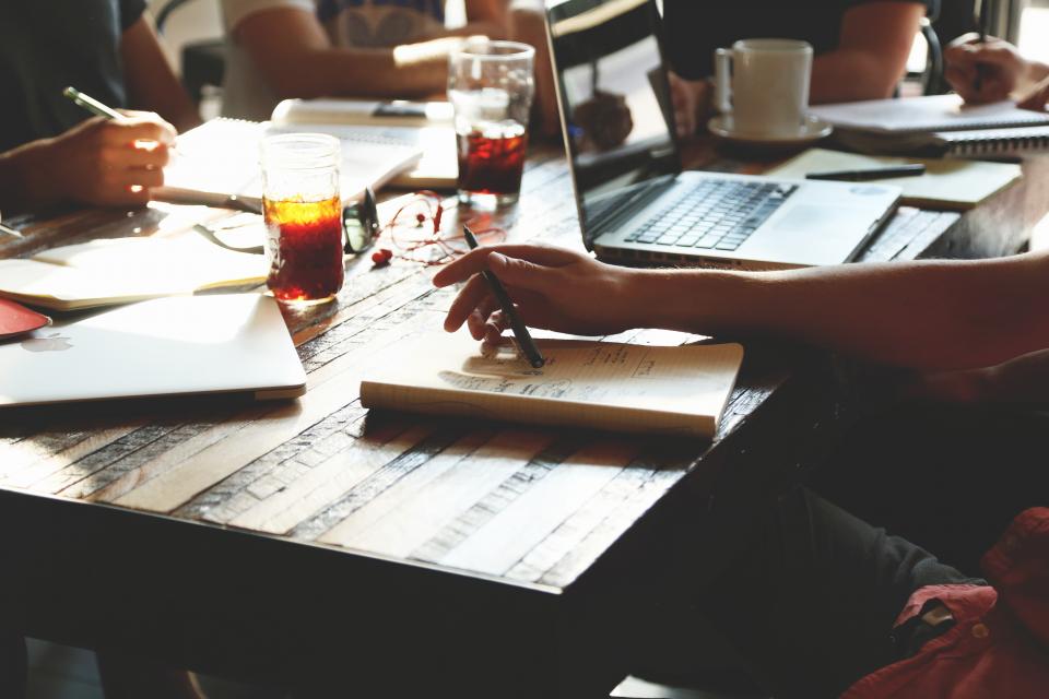 Menschen sitzen mit Unterlagen und Laptops an einem Holztisch und besprechen sich.